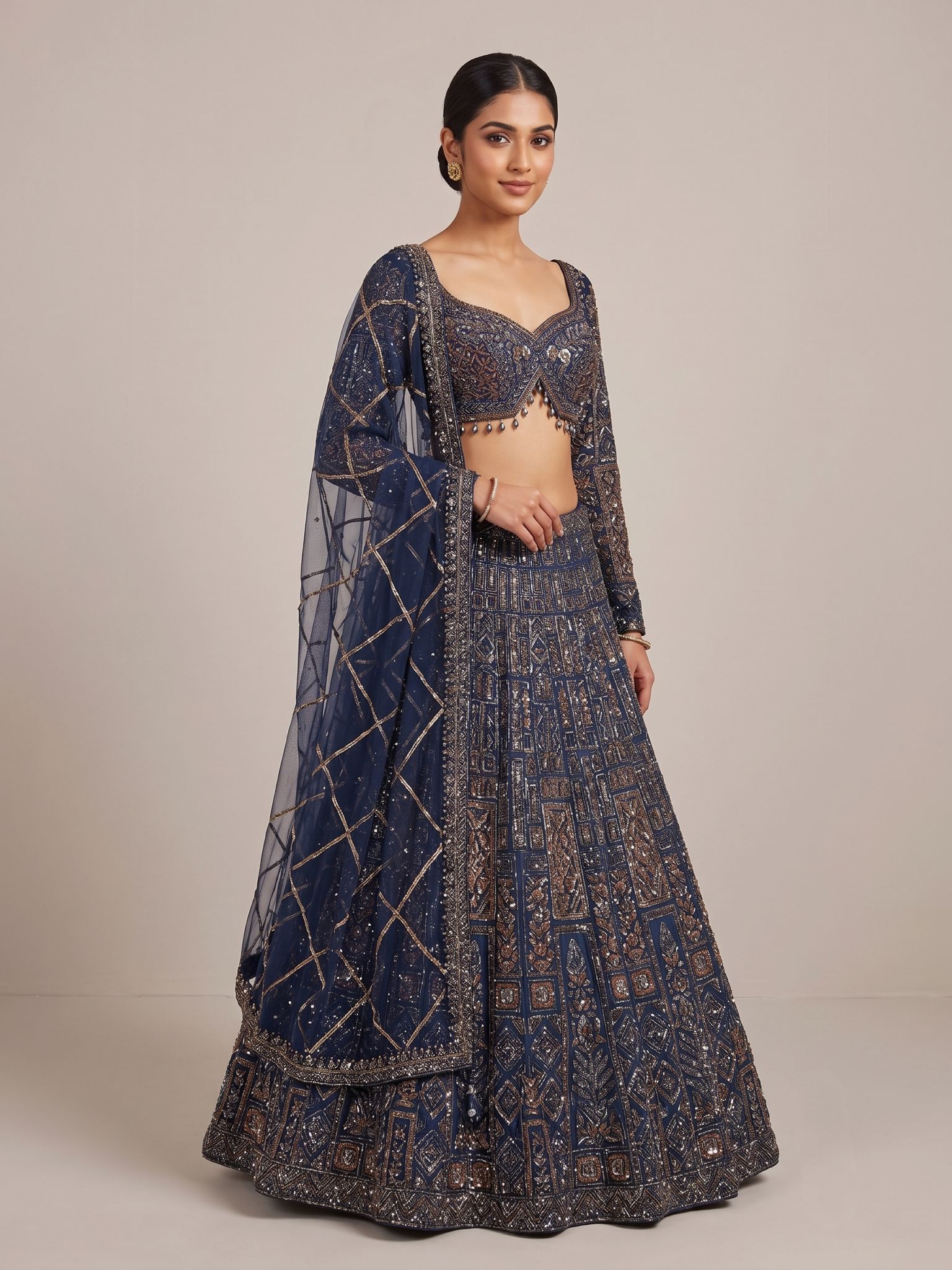 A Yashwanti London women's bridal lehenga choli in a deep navy-blue base, detailed with antique copper and gold embroidery in geometric and floral motifs accented with sequins and bead work. The fitted choli has a sweetheart neckline, full sleeves, and a deep open back fastened with a tie-up dori and beaded latkan drops. The flared A-line skirt carries dense panelled embroidery that builds into a broad ornamental hem, paired with a sheer net dupatta finished in diamond-lattice gota detailing.